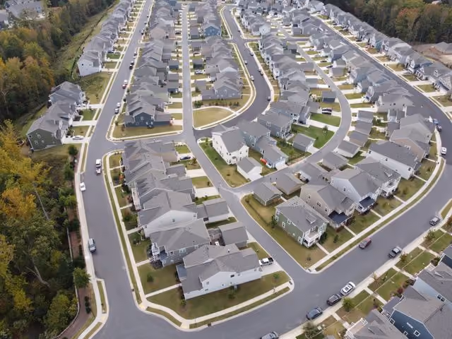 Aerial view of a suburban neighborhood showing rows of similar detached houses along curved streets with green lawns and some parked cars.