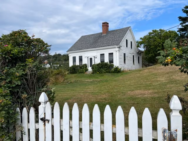 White traditional house with a steep roof and chimney on a grassy hill, viewed through an open white picket gate with greenery on the sides under a partly cloudy sky.