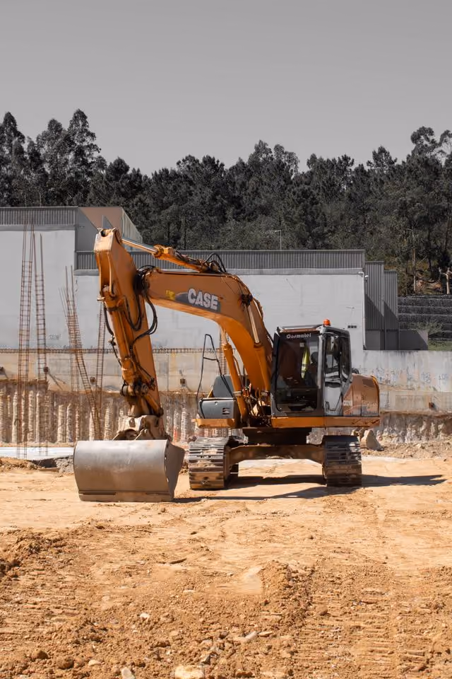 Orange CASE excavator with bucket attachment on a dirt construction site with a partially built structure and trees in the background.
