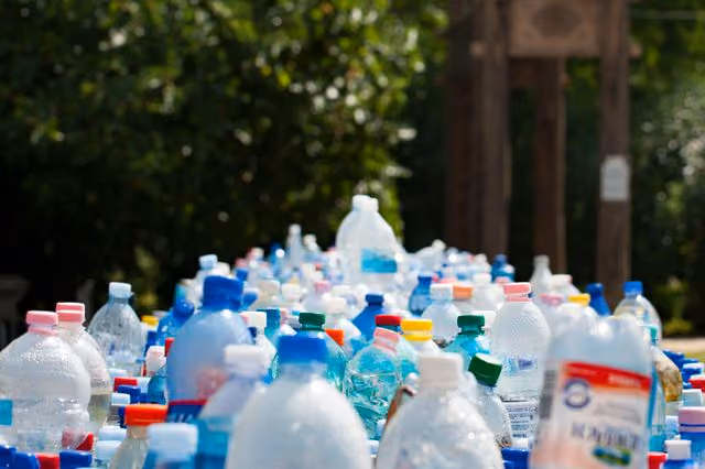 A large collection of various empty plastic water bottles with different colored caps grouped together outdoors, with blurred greenery and a wooden structure in the background.