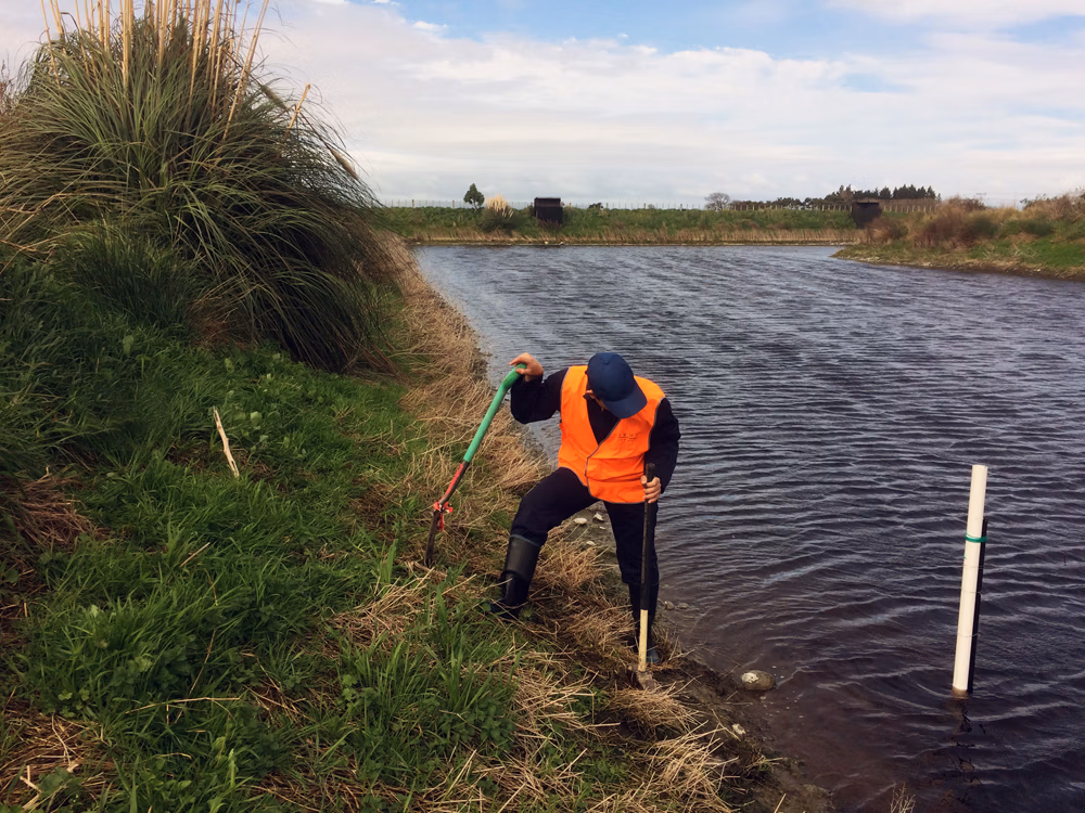 Person wearing an orange safety vest, black pants, and rubber boots standing by a grassy bank next to a body of water, using a green-handled tool for fieldwork or environmental survey.