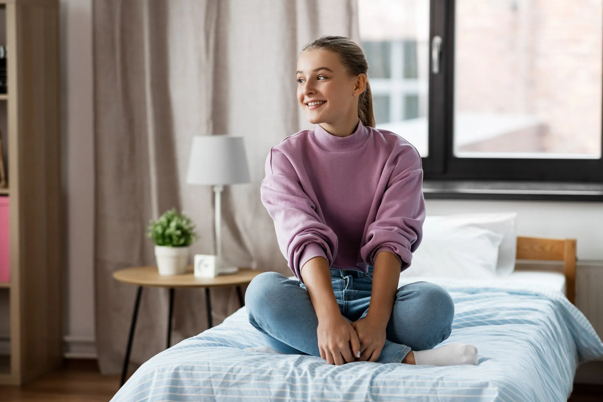 Smiling young woman in a purple sweater sitting cross-legged on a bed in a cozy room.