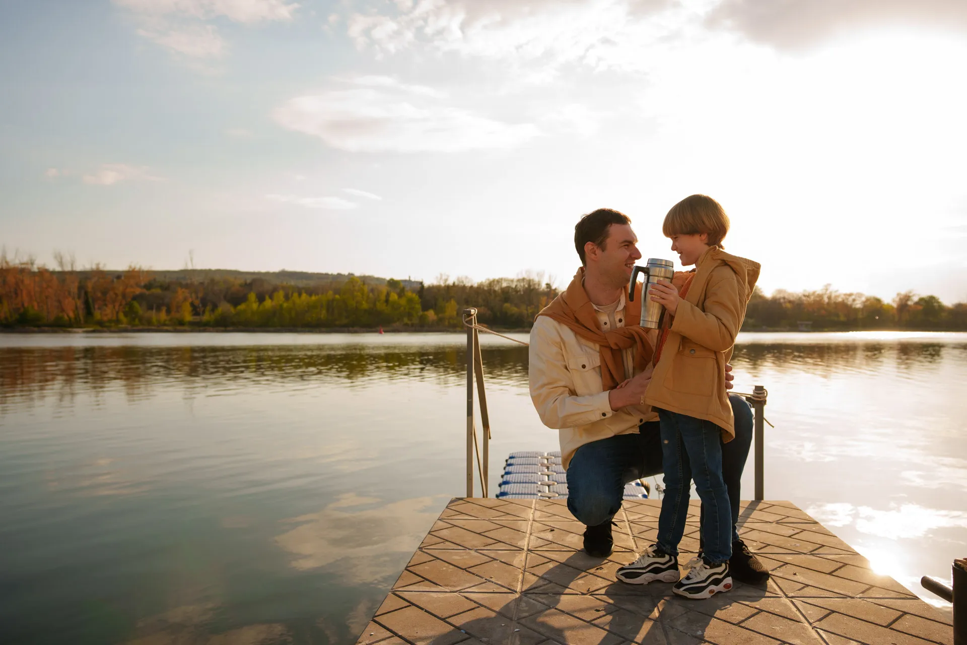 Father kneeling on dock, smiling at son holding a travel mug by the lake at sunset.