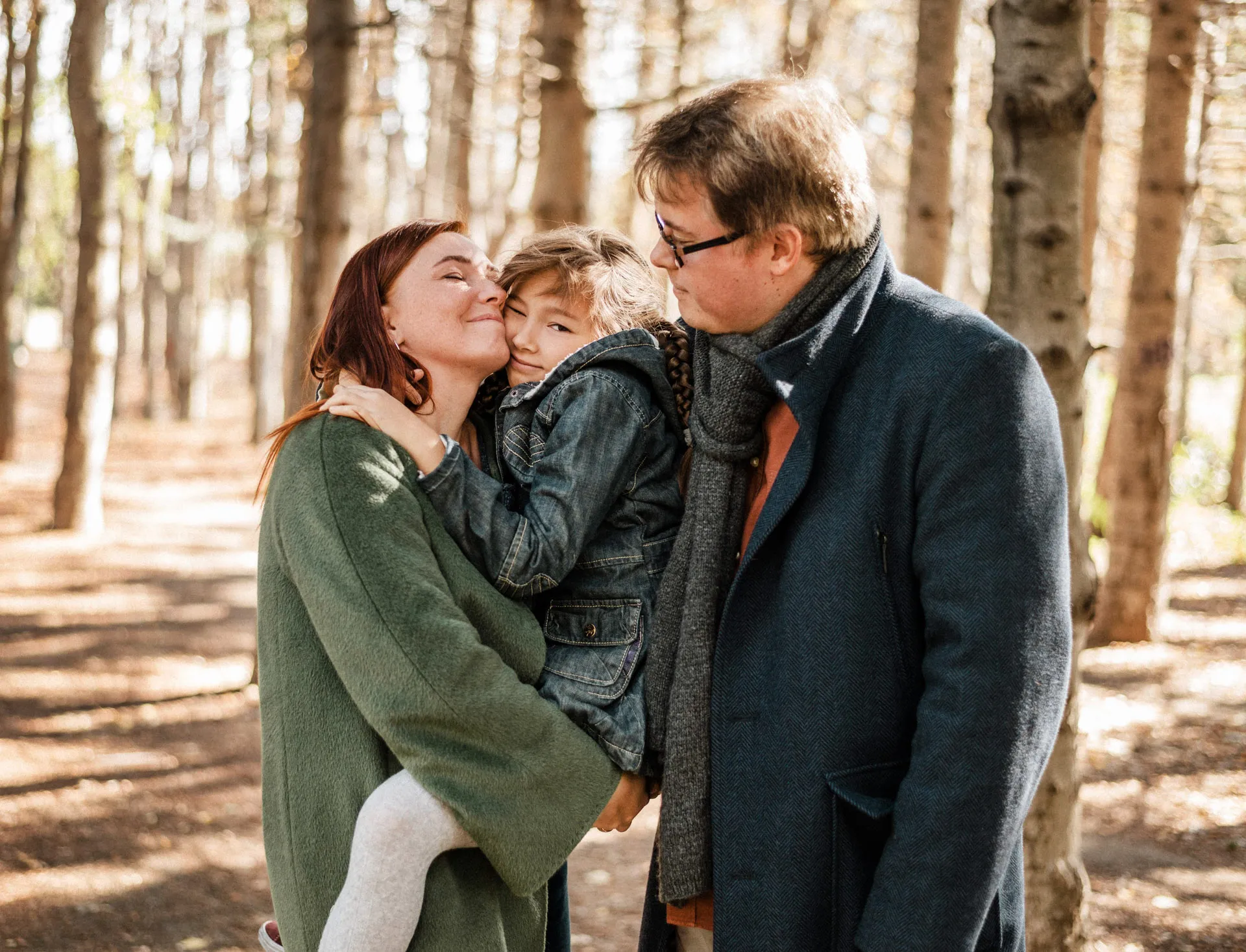 Parents outdoors in autumn woods, mother hugs and kisses daughter, father watches smiling.
