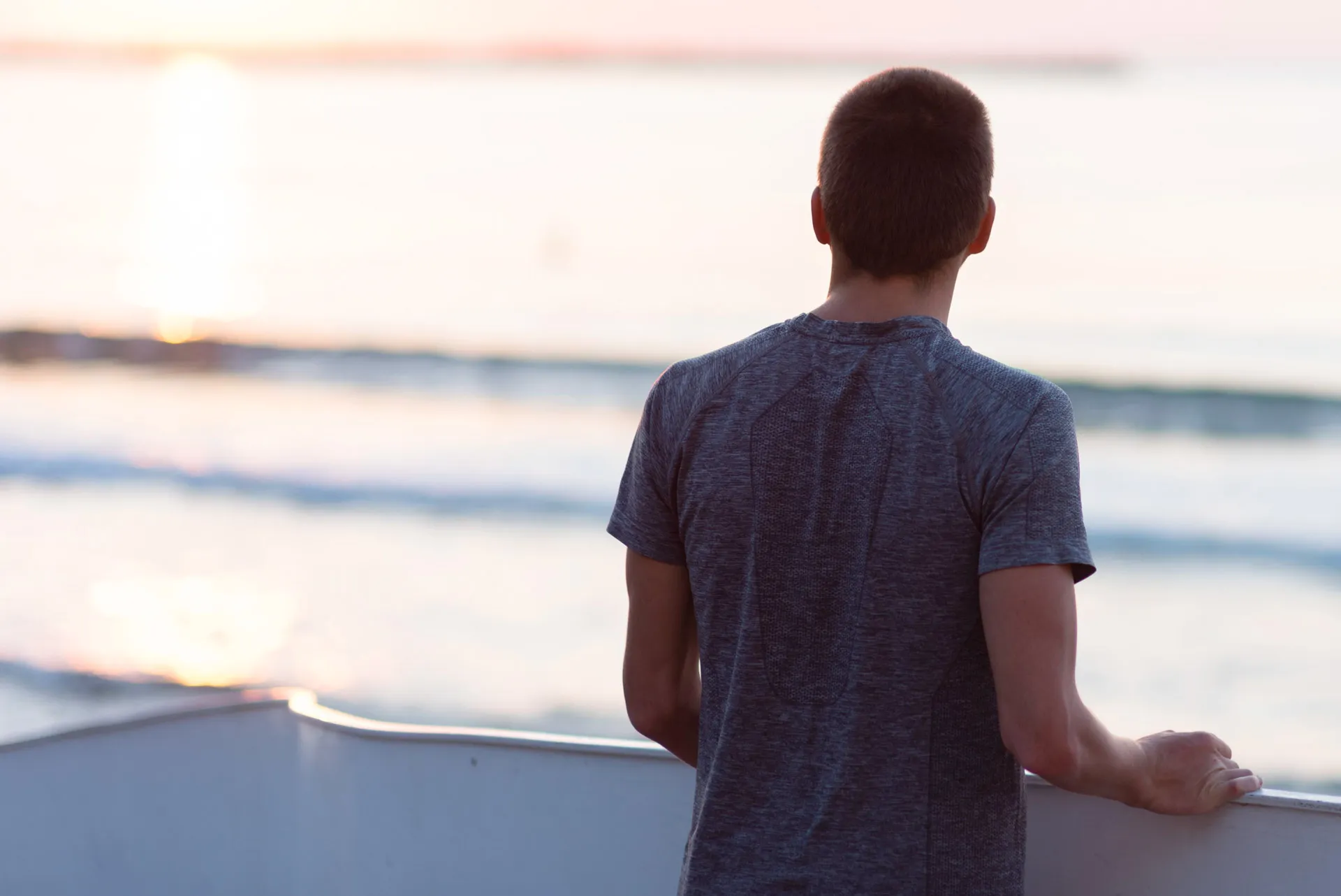 Young man in a gray shirt standing on a boat, looking at the calm sea during sunrise or sunset.