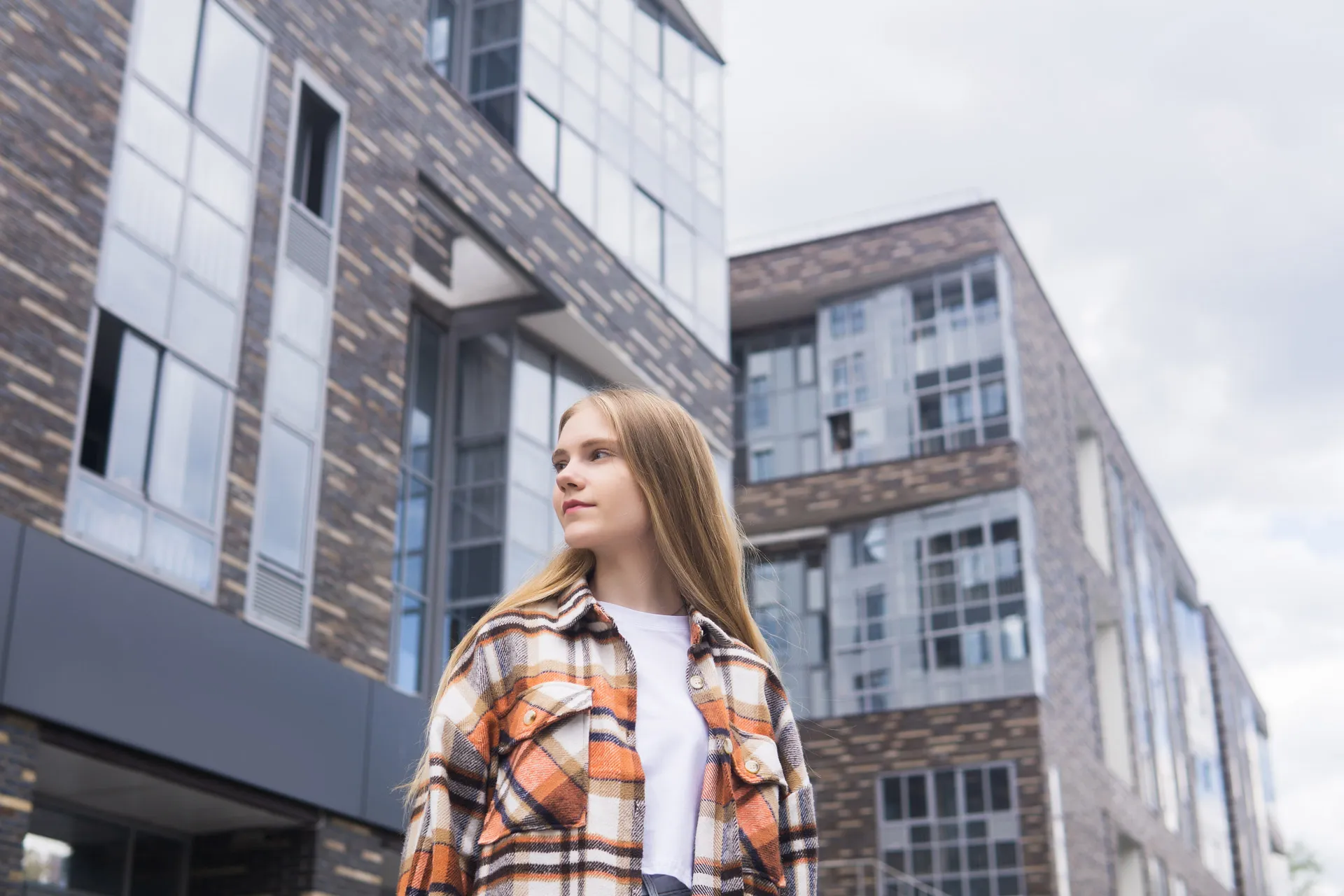 Young woman with long blonde hair wearing an orange plaid shirt standing in front of modern buildings.