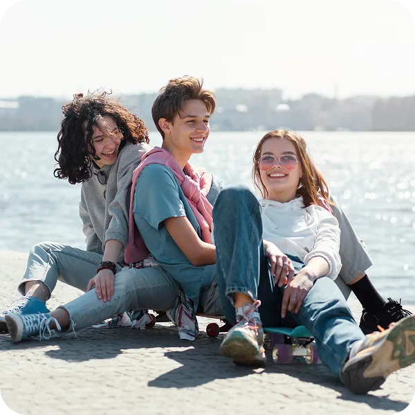 Three teens sitting and smiling by the waterfront in casual clothes with a skateboard.