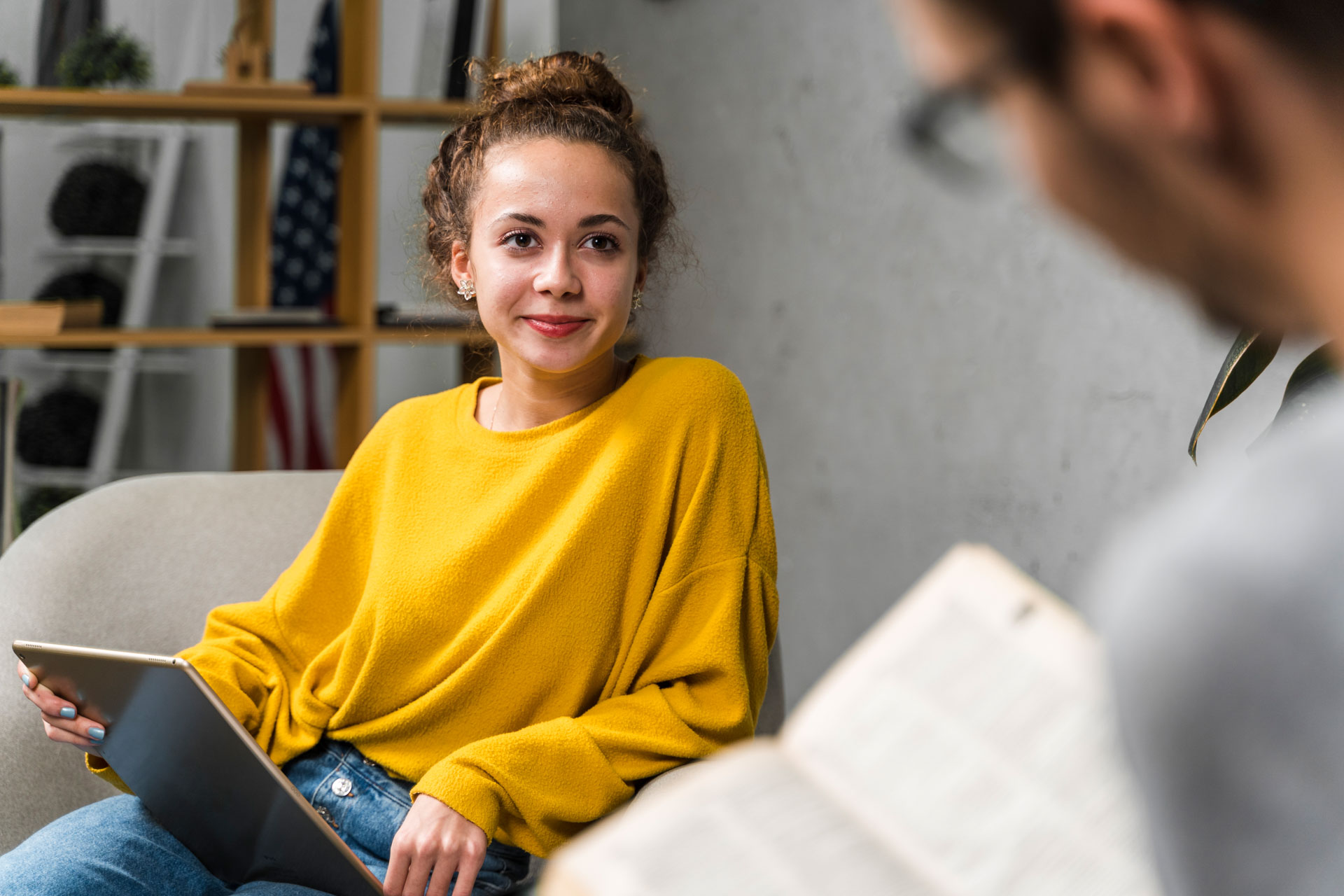 Young woman in yellow sweater holding a tablet, looking at a blurred person reading a book.