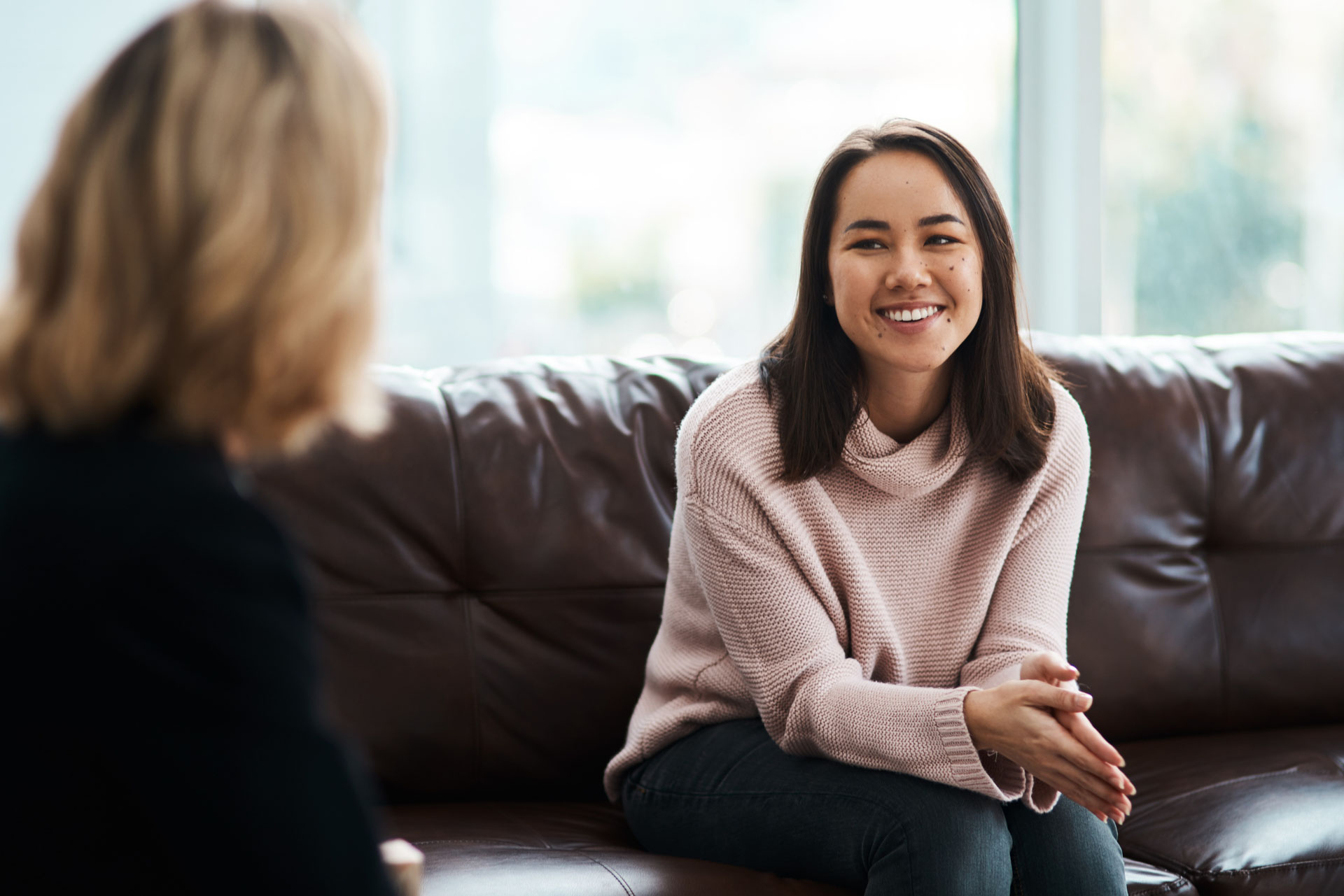 Smiling woman in a pink sweater sitting on a couch talking to another person.