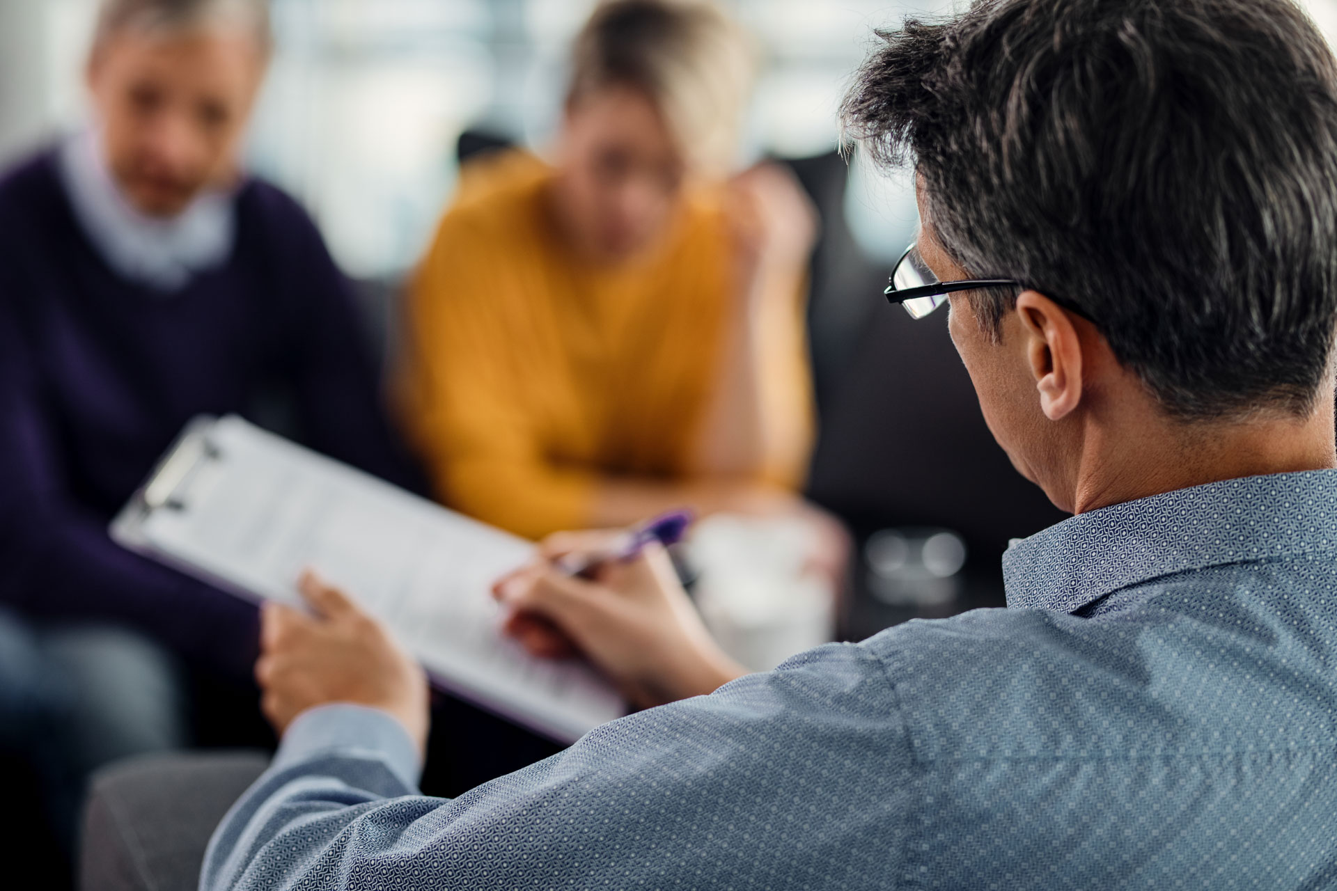 Therapist with glasses taking notes on clipboard while couple sits closely in background.