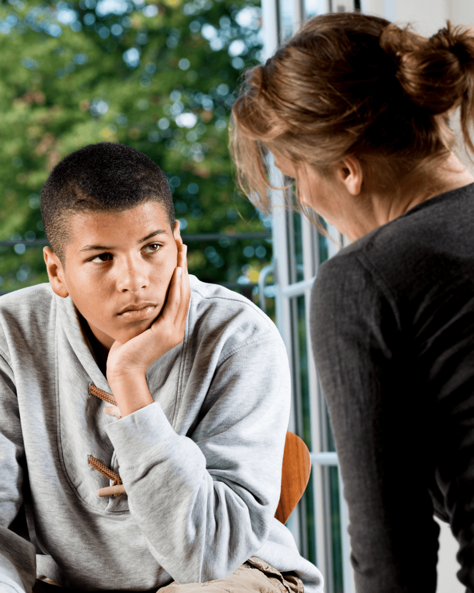 Teen boy with serious expression resting his face on his hand, talking to a woman indoors.