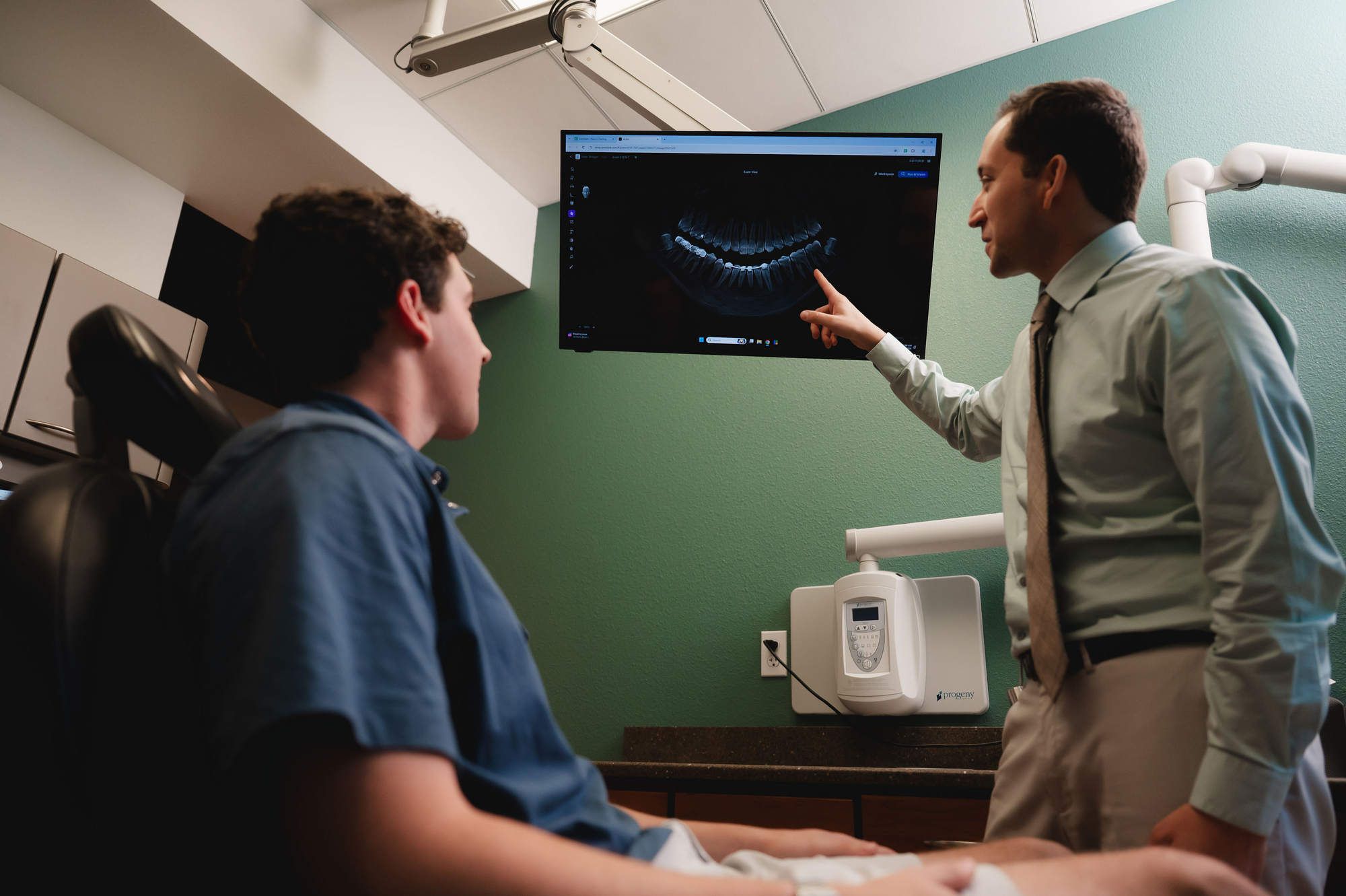 Dentist pointing to a dental X-ray on a screen while a patient sits in the exam chair.