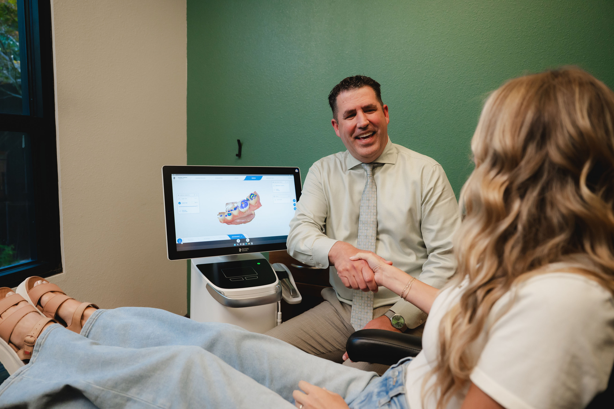 Dentist shaking hands with a female patient sitting in a dental chair beside a dental scan monitor.