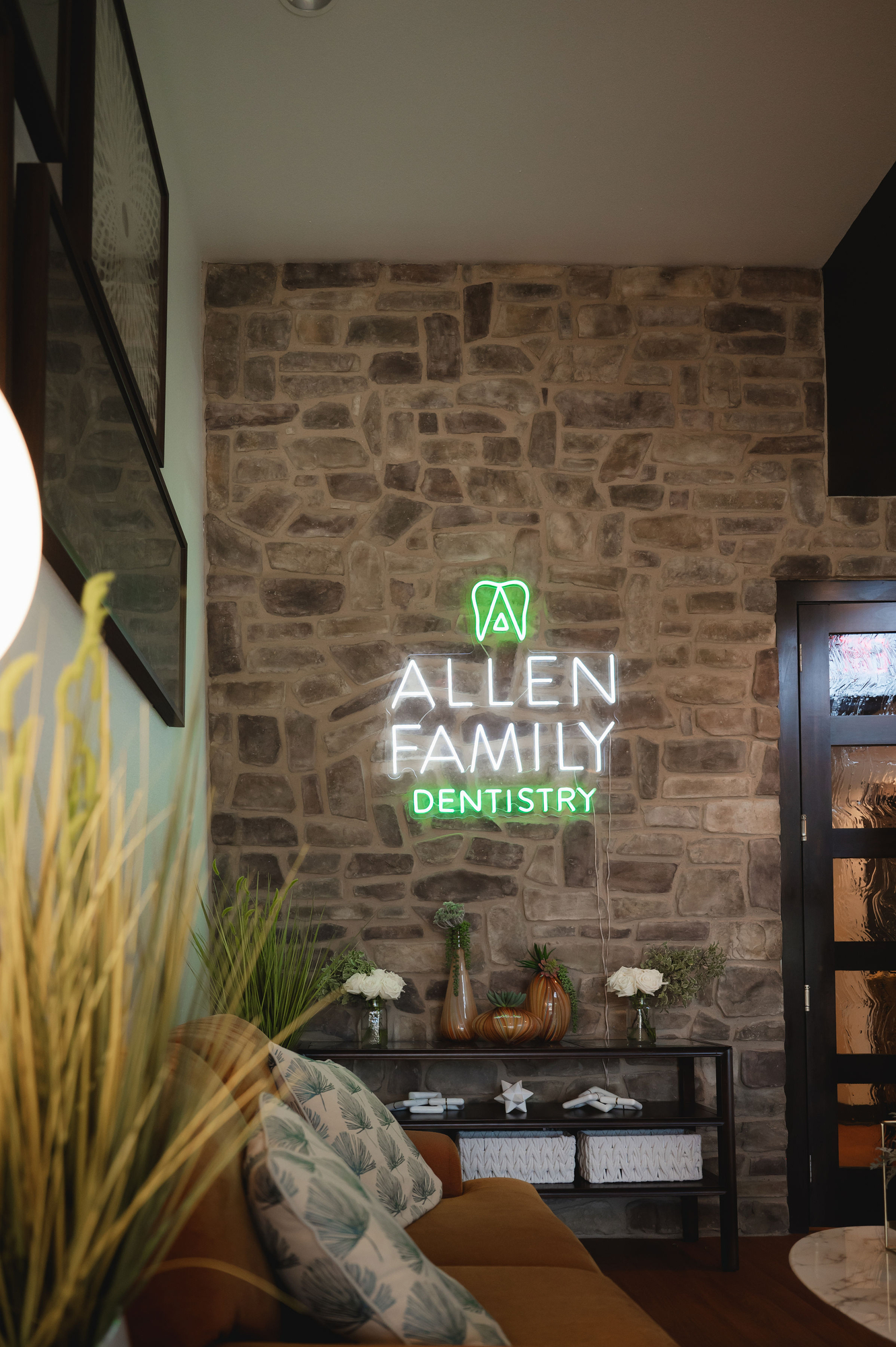 Cozy waiting area with brown couch, green plants, and neon sign reading Allen Family Dentistry on stone wall.