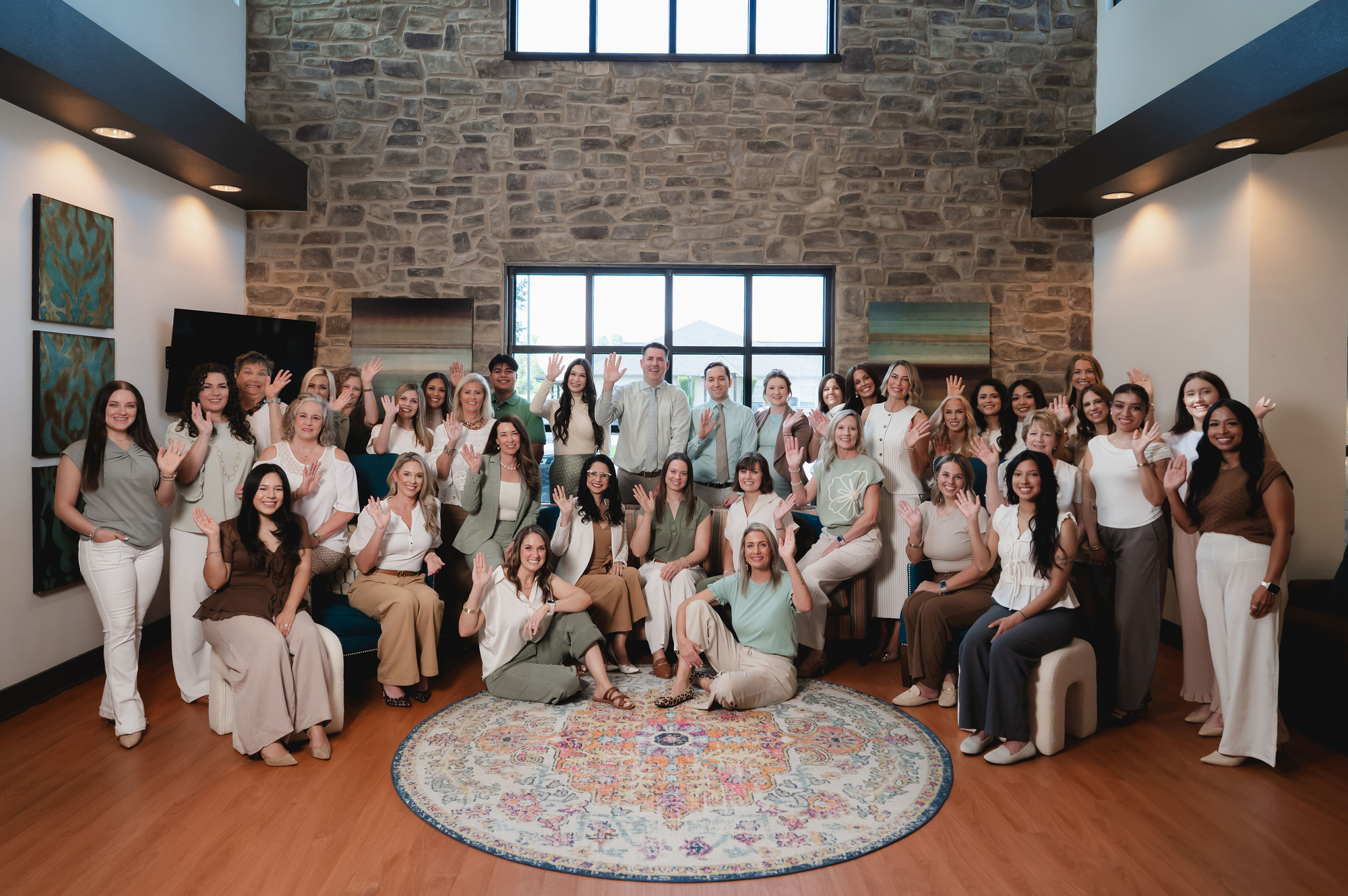 Large group of diverse people waving inside a room with stone walls and large windows.