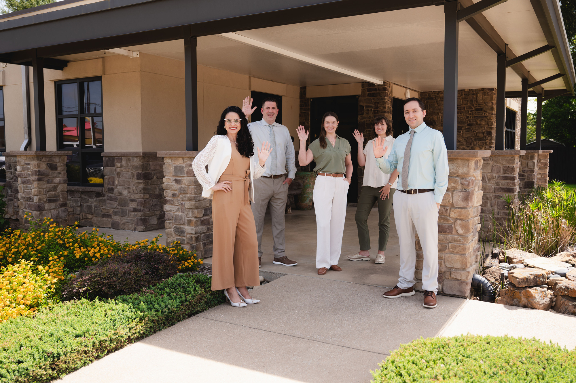 Five professionals waving while standing under a covered walkway at a stone building entrance.