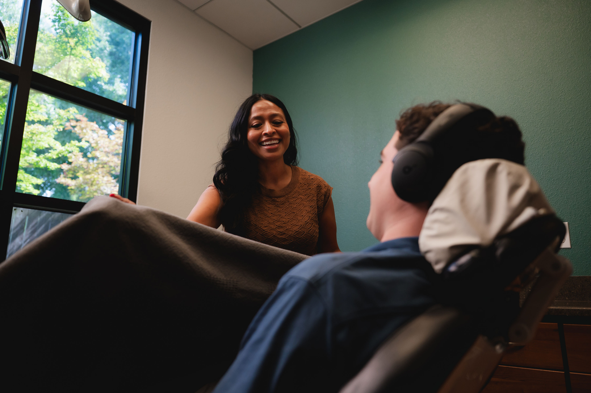 Woman smiling at a relaxed patient wearing headphones sitting in a dental chair by a window.