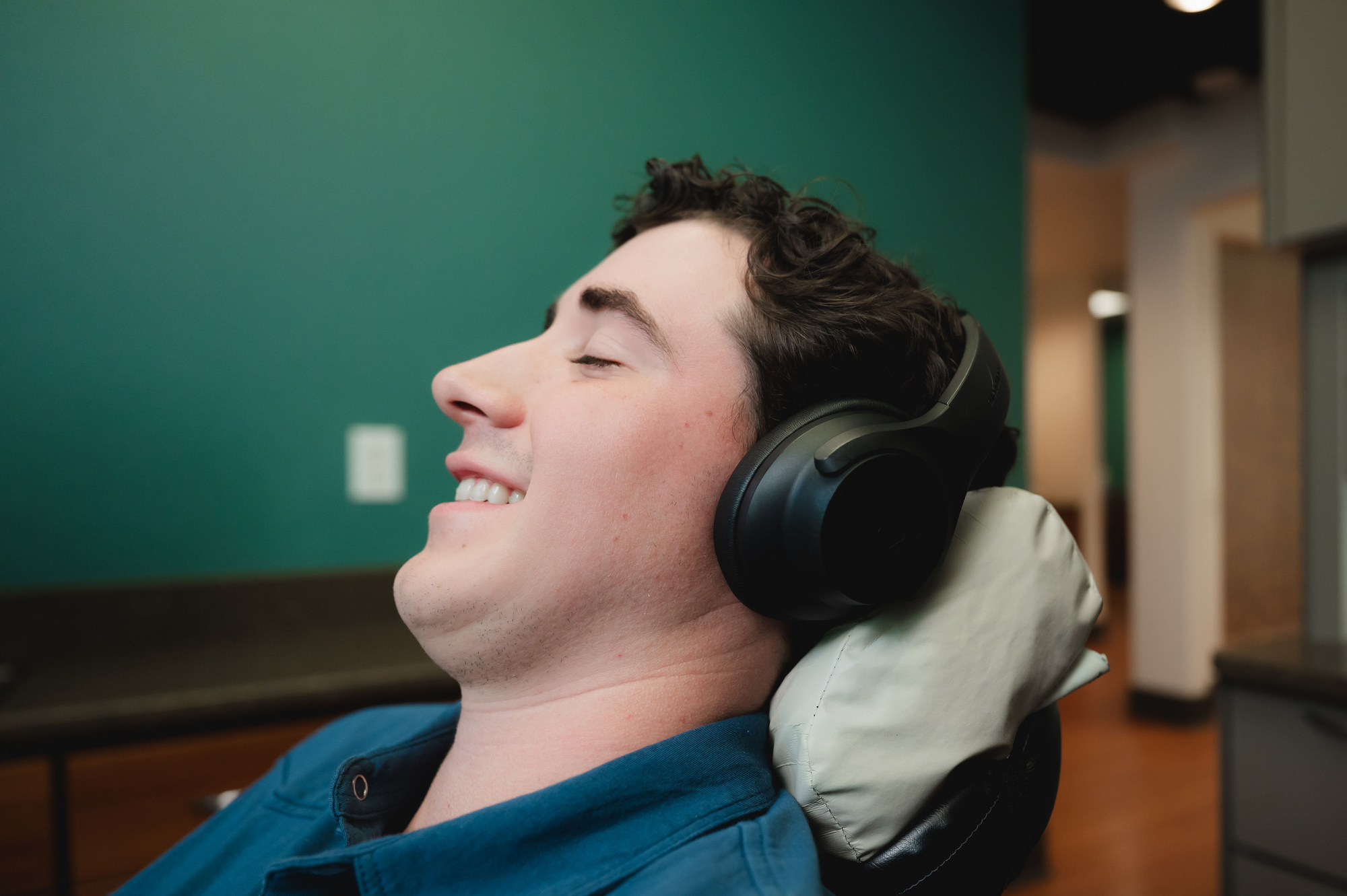 Young man with closed eyes wearing black headphones, reclining and smiling in a room with green wall.