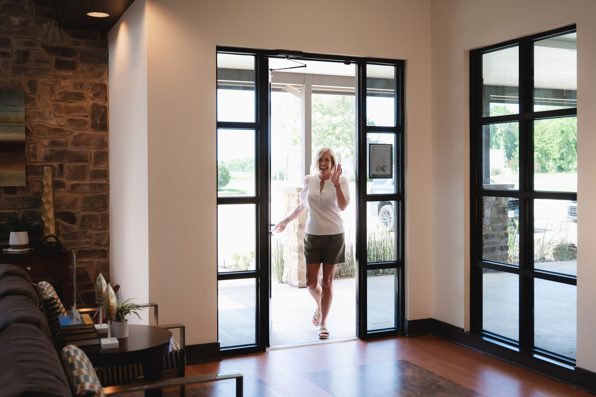Woman entering a bright room through glass double doors, smiling and waving.