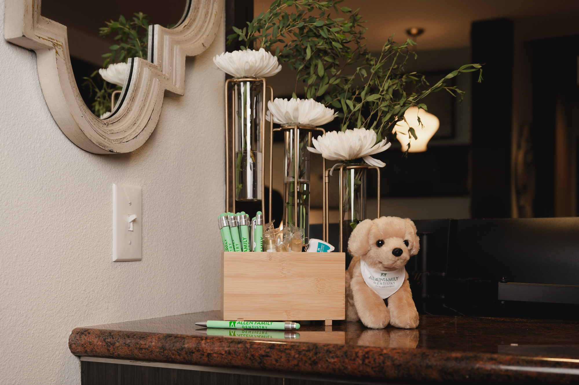 Plush dog wearing Allen Family Dentistry bandana next to a wooden box with pens and flowers in vases on counter.