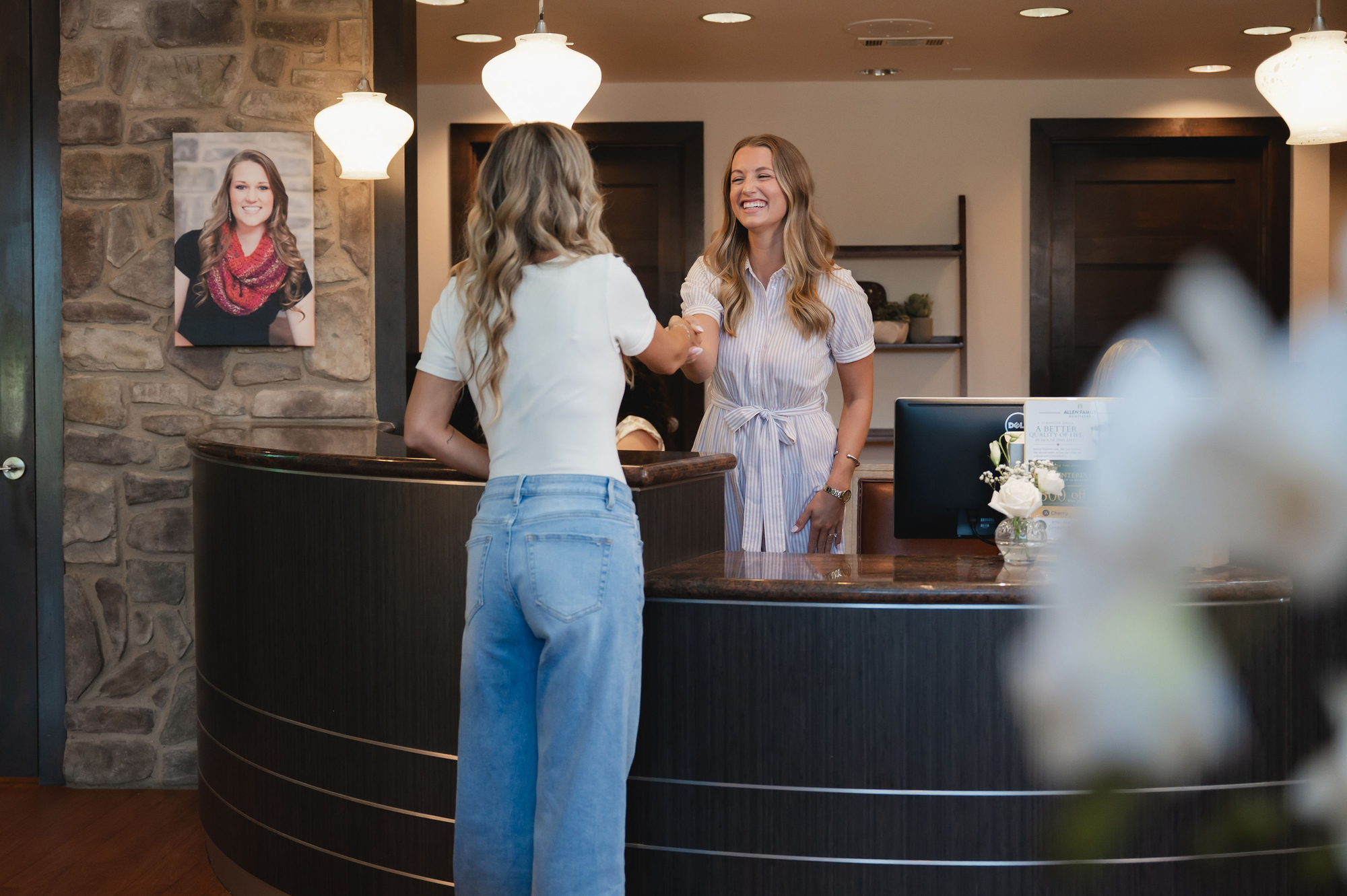 Woman in white shirt shaking hands with receptionist in striped dress at curved wood front desk.