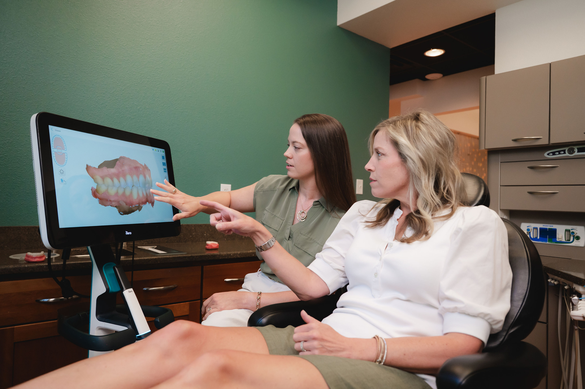 Two women view and discuss 3D dental teeth model on a screen in a dental office.
