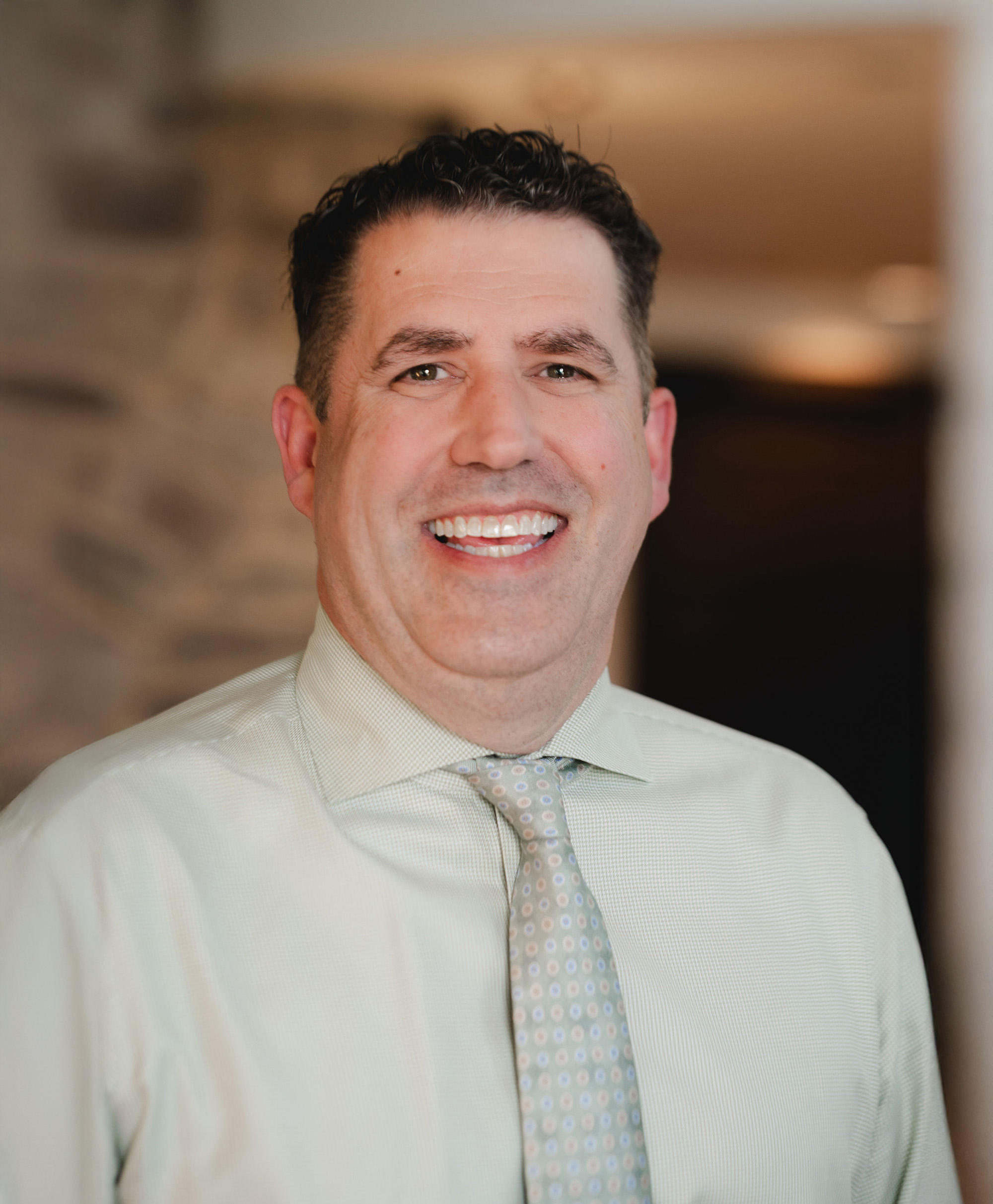 Smiling man wearing a light green shirt and patterned tie against a blurred background.