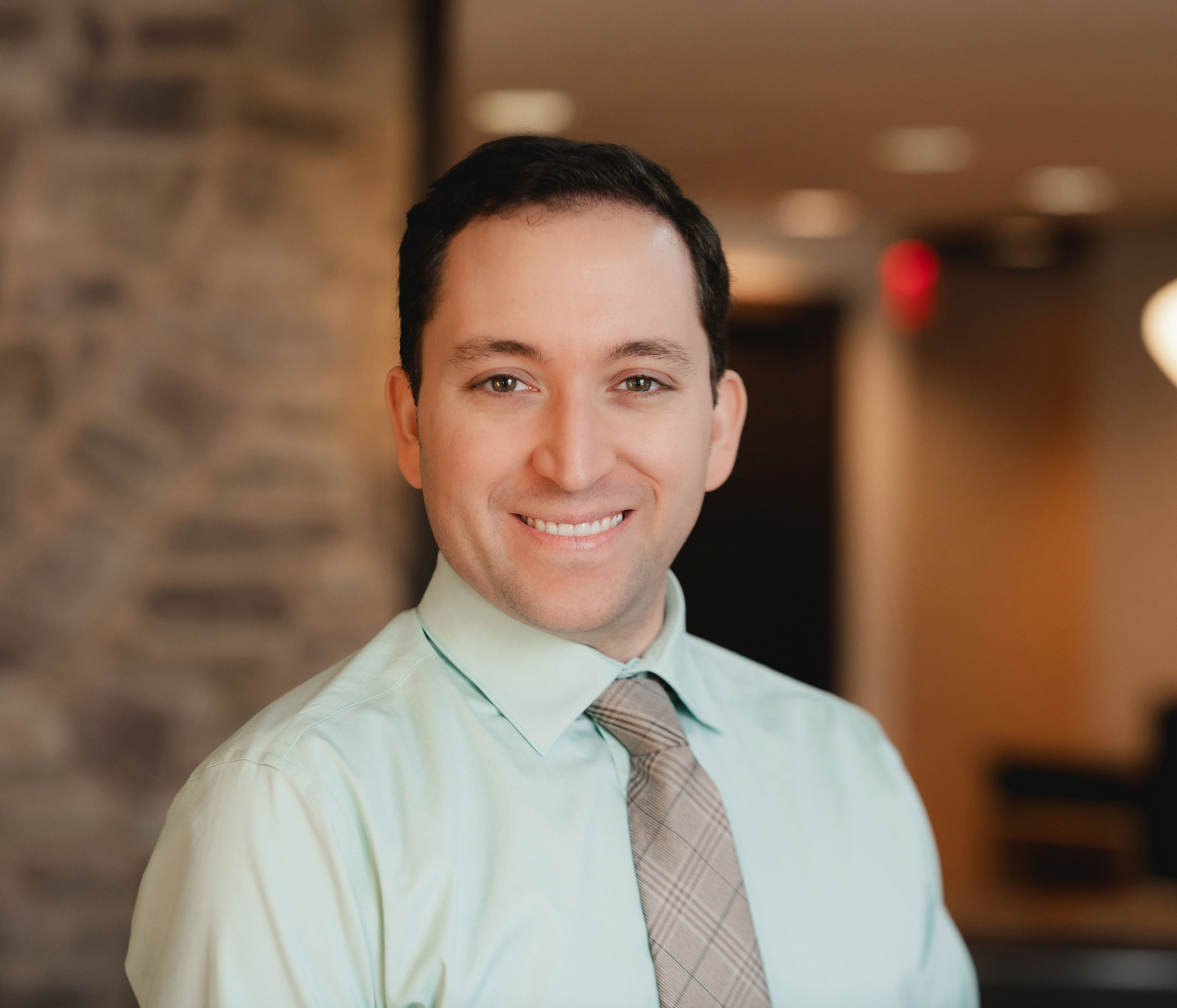 Smiling man wearing light green shirt and plaid tie standing indoors with a stone wall background.