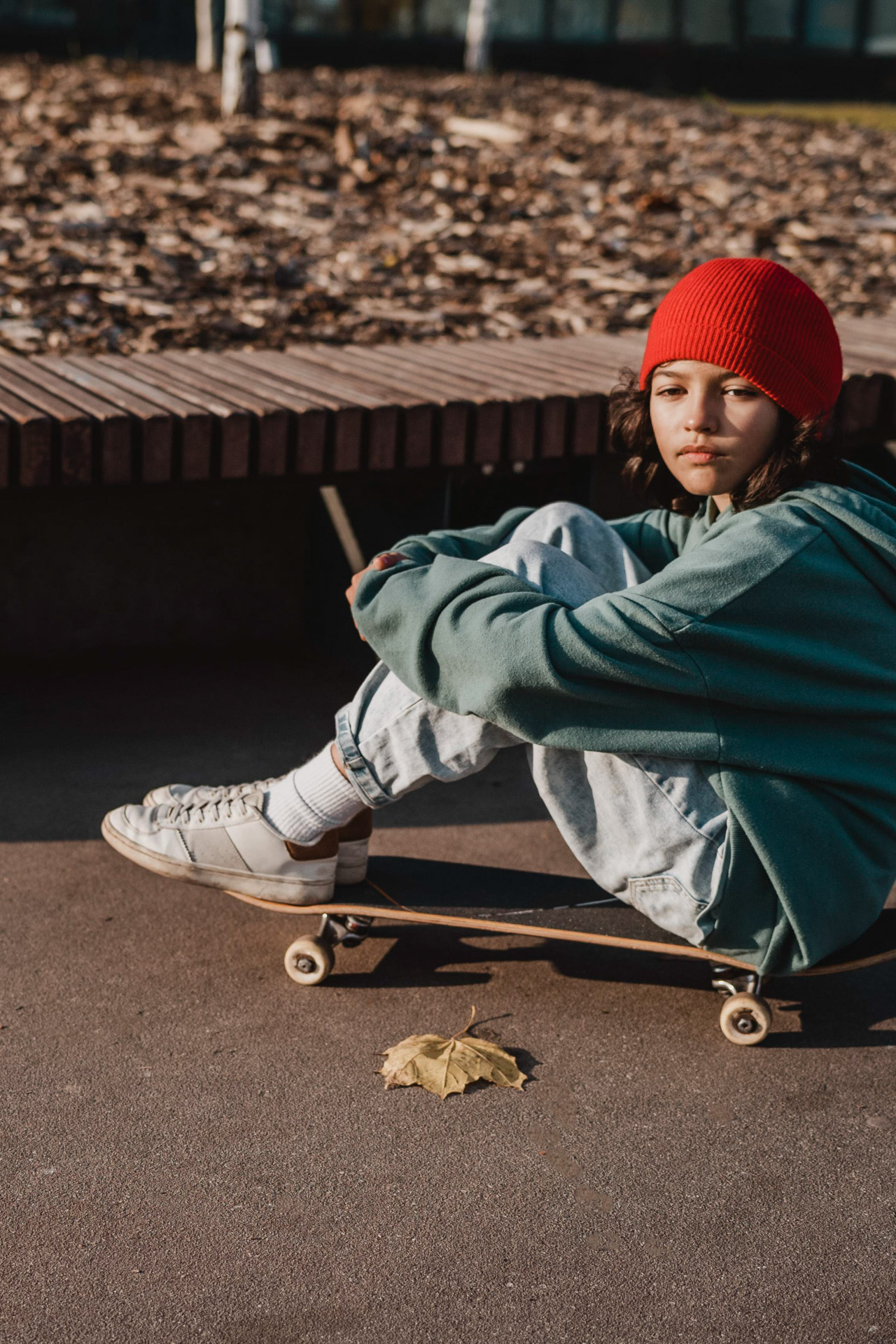 Teenager in red beanie sitting on skateboard on pavement near bench during daytime.