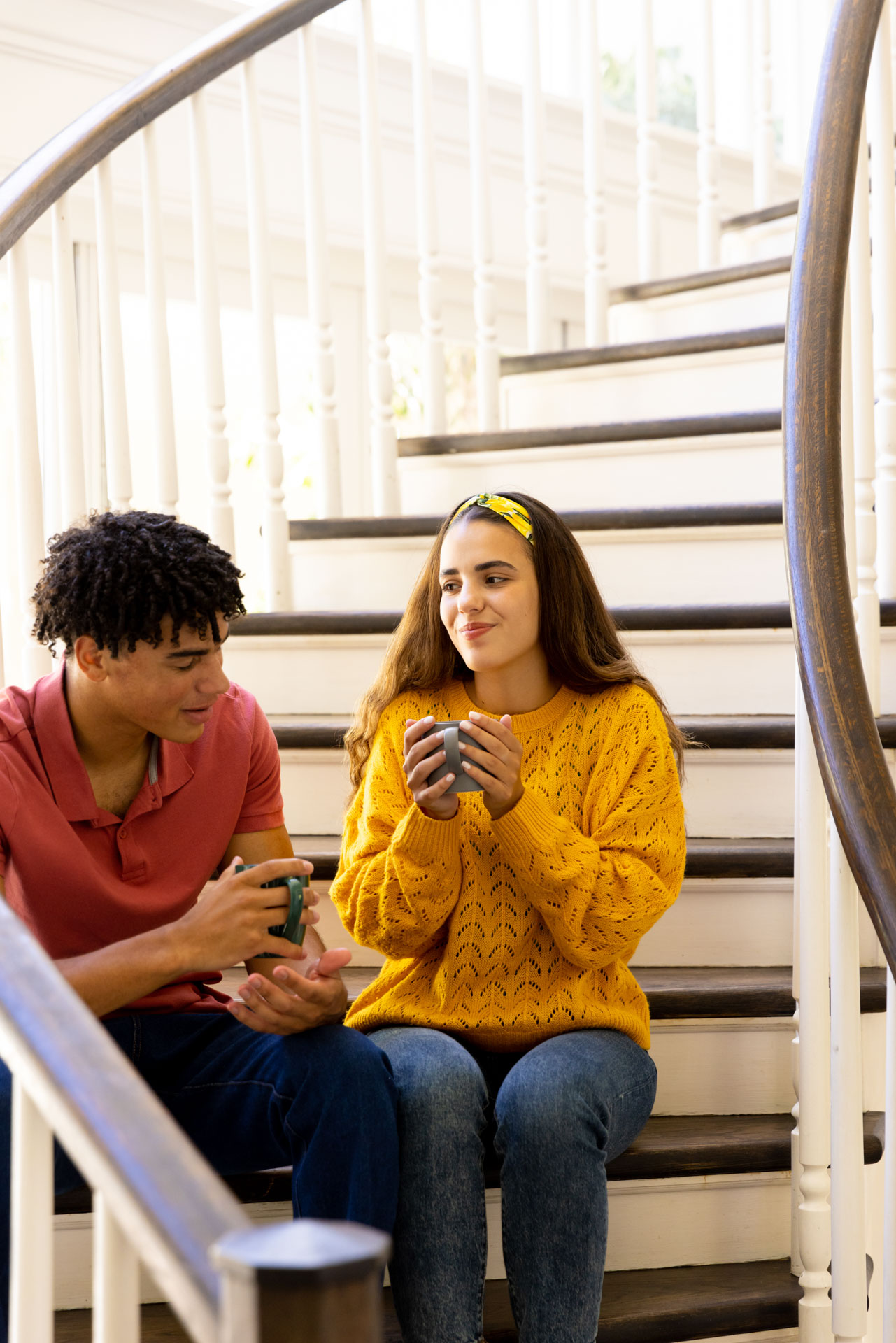 Young couple sitting on stairs indoors, each holding a coffee cup, engaged in conversation.