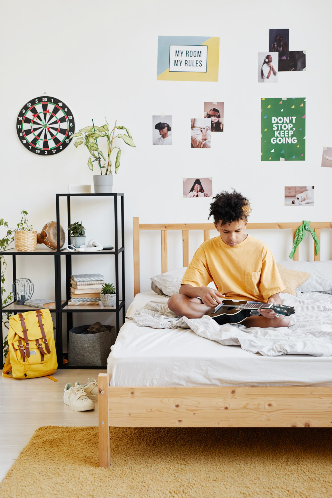Teenage boy in yellow shirt sitting cross-legged on bed playing guitar in a decorated bedroom.