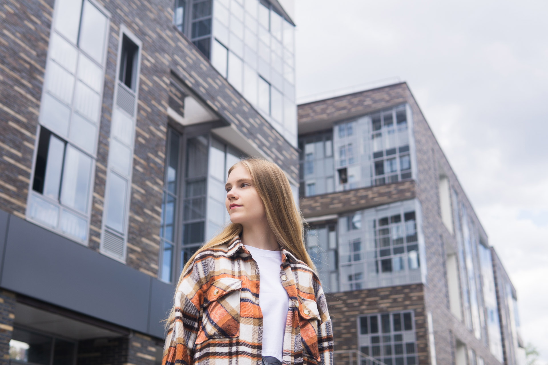 Young woman with long blonde hair wearing an orange plaid shirt standing in front of modern buildings.