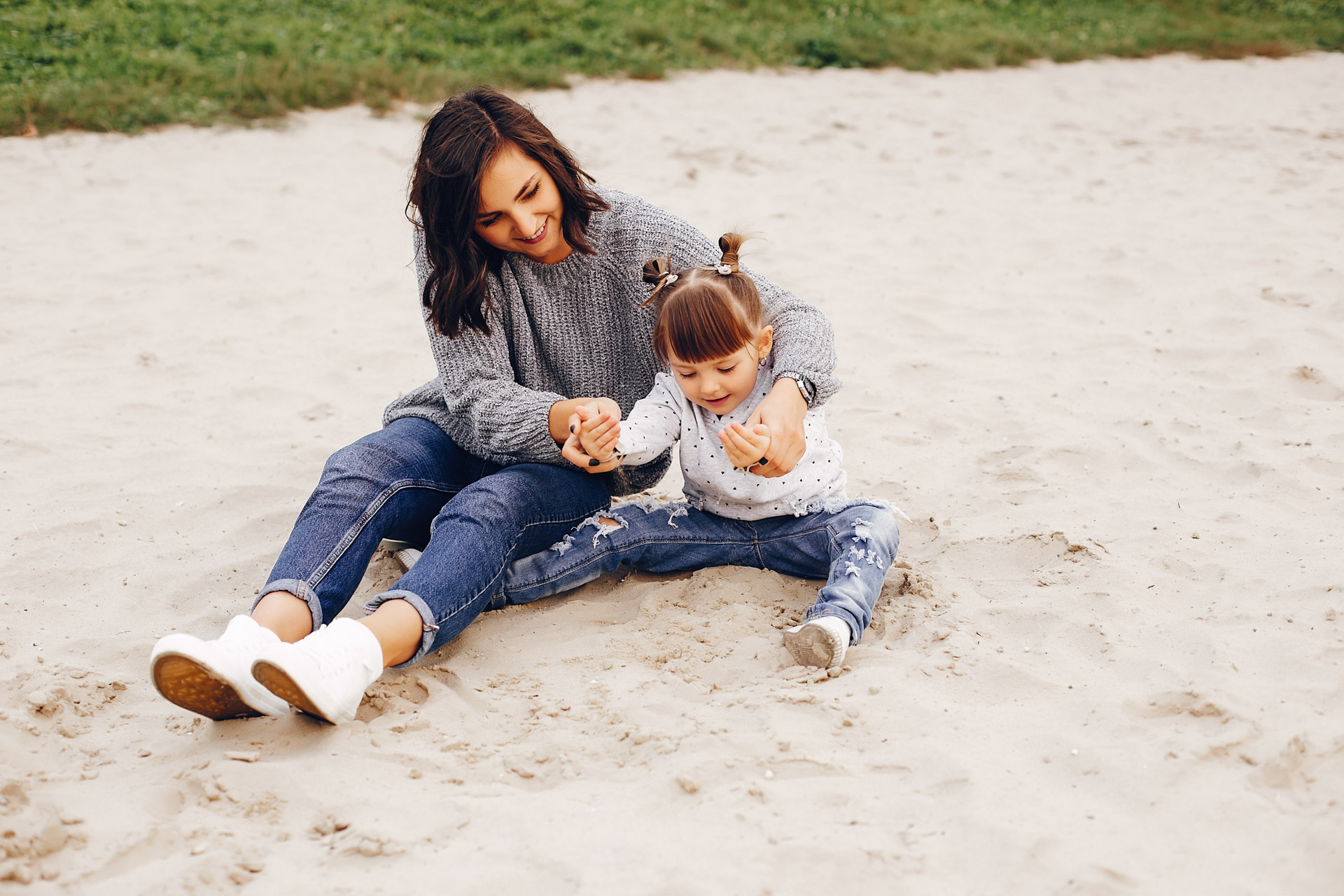 Mother and daughter sitting in sand, playing and smiling together in a park.
