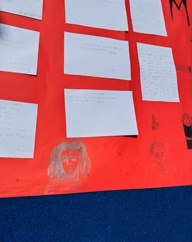 Students display written work and drawings on a red poster board