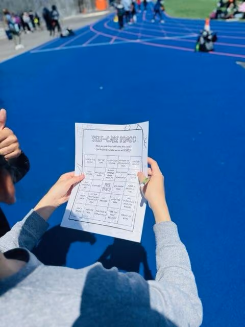 Student holding a self-care bingo activity sheet at a track event