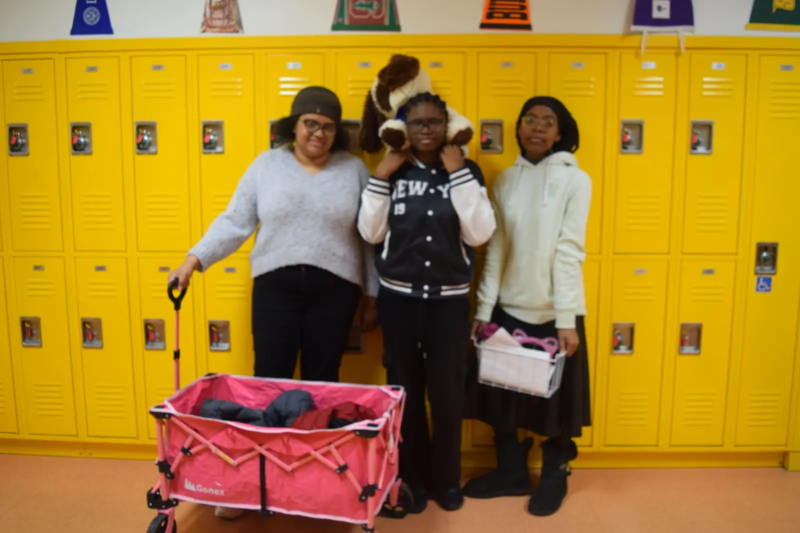 Three students posing together by yellow school lockers with donation cart