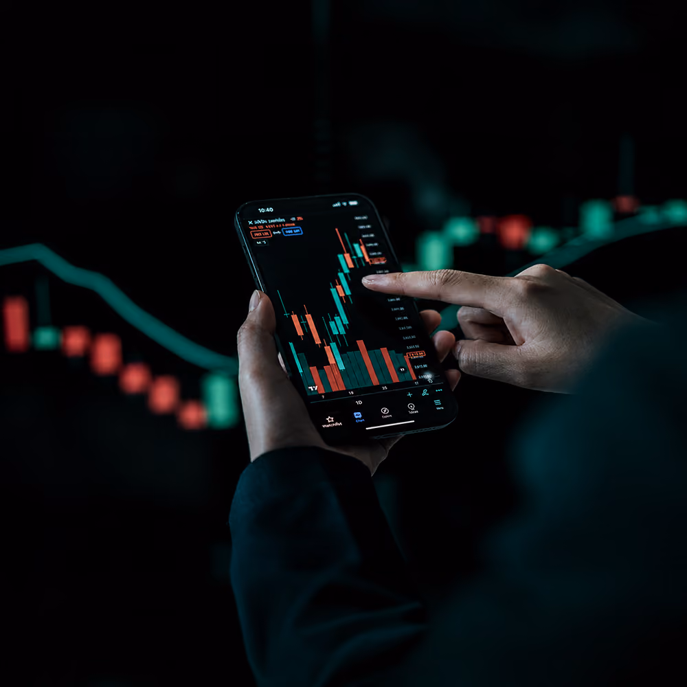 Person using a smartphone showing red and green candlestick stock charts on a dark background.