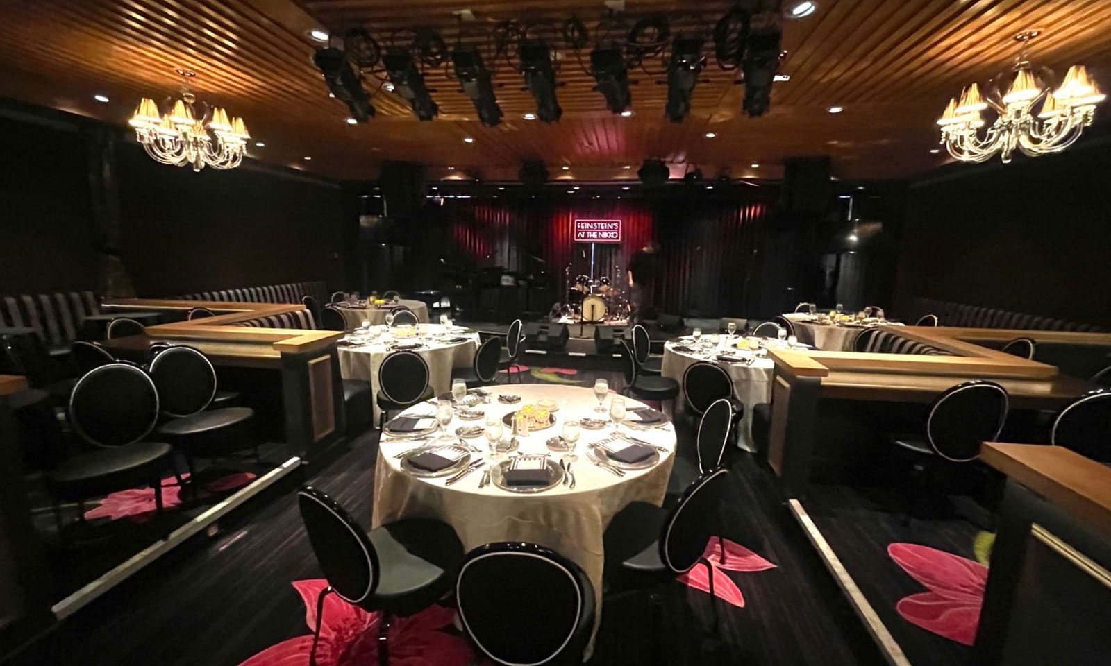 Elegant supper club dining room with round, formally set tables facing a small stage with a drum kit and Feinstein’s at the Nikko sign.