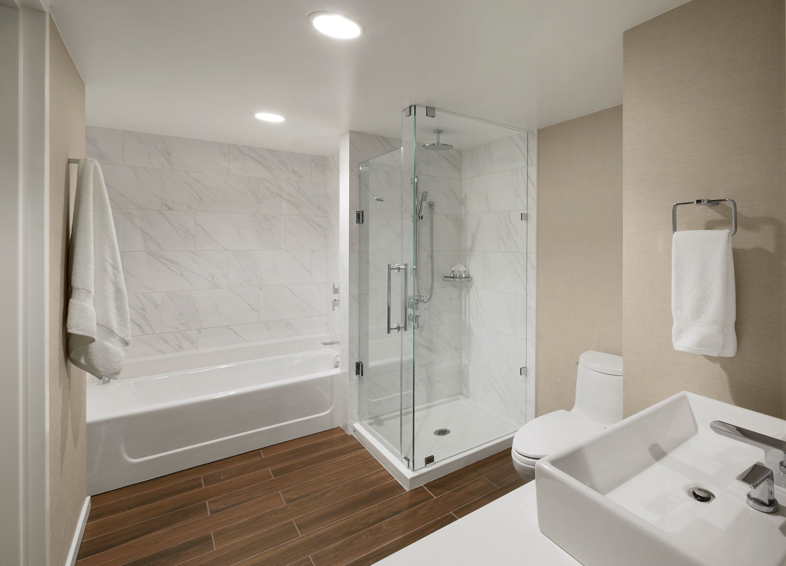 Hotel bathroom with a soaking tub, glass-enclosed marble shower, wood-look flooring, toilet, and white vessel sink.