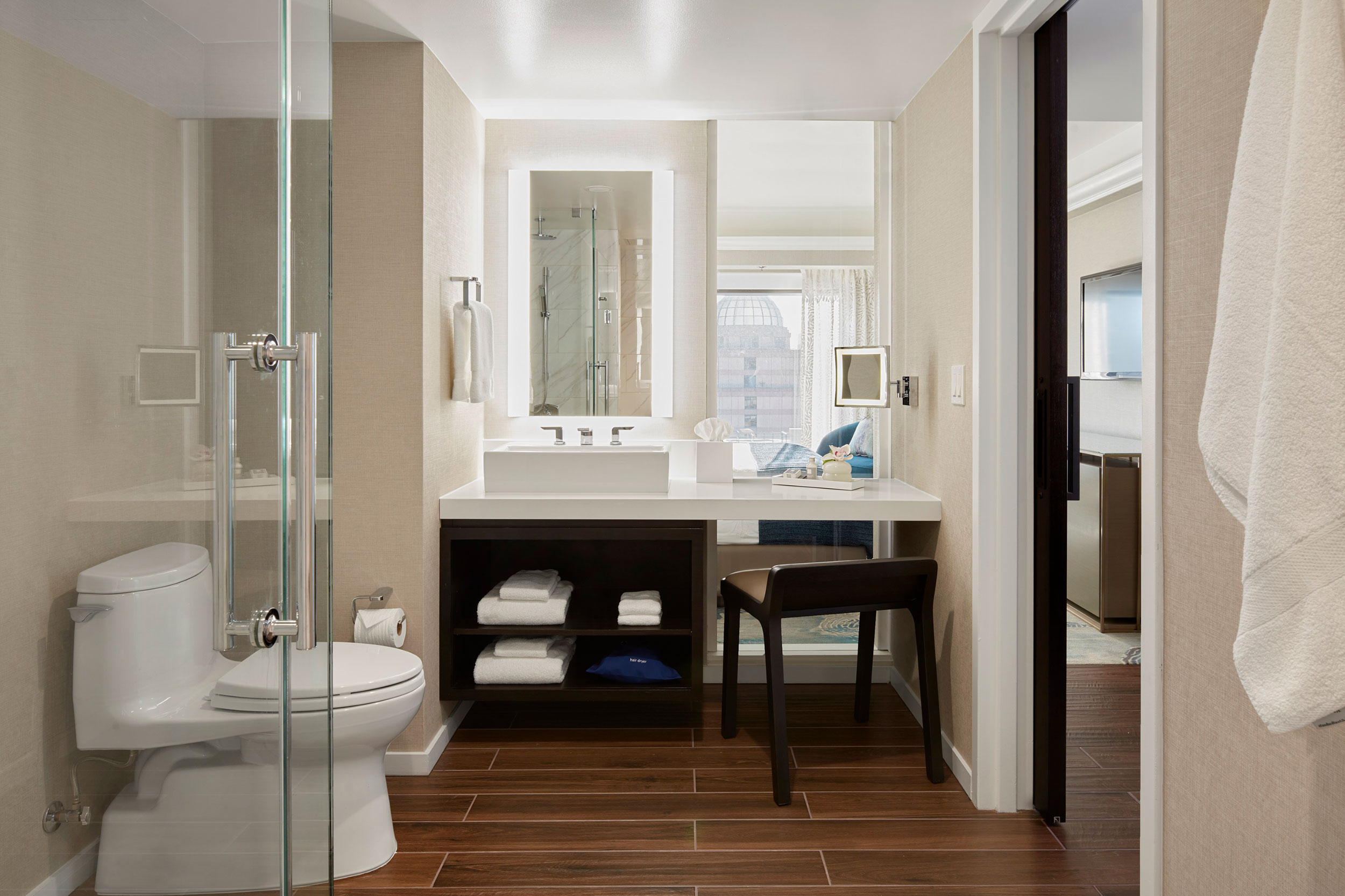 Modern hotel bathroom with a glass shower, toilet, illuminated vanity mirror, vessel sink, and dark wood cabinetry.
