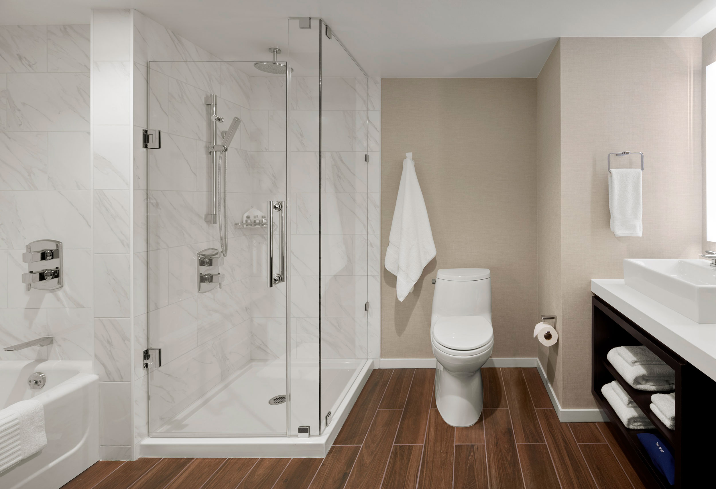 Spacious hotel bathroom with a marble-tiled glass shower, soaking tub, toilet, vessel sink, and wood-look flooring.