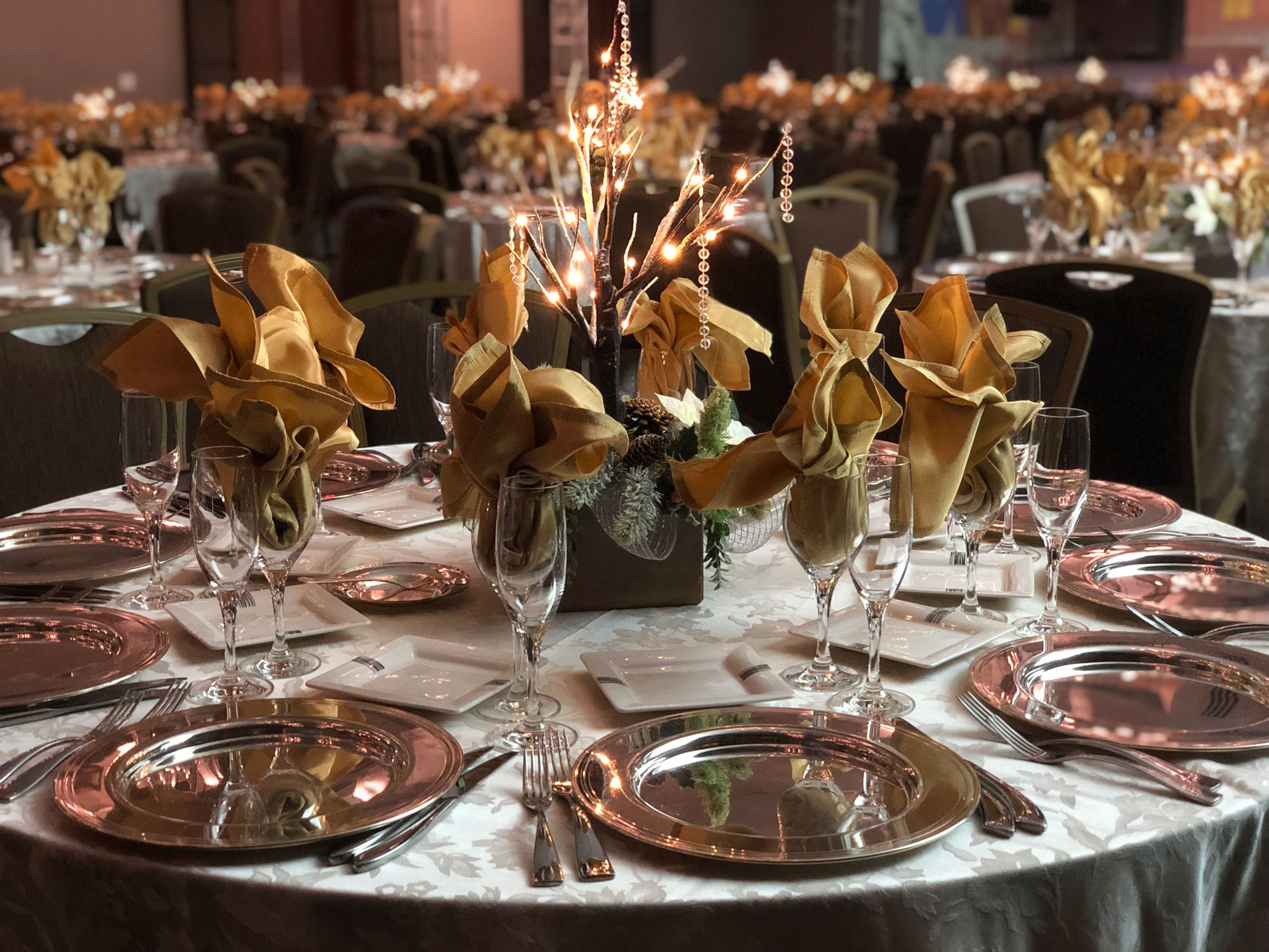 Round banquet table with gold napkins, metallic chargers, and illuminated holiday centerpiece.