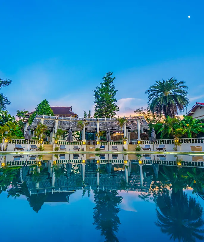 Outdoor pool area at dusk with a clear blue sky, palm trees, and a building with a pergola reflected in the water.