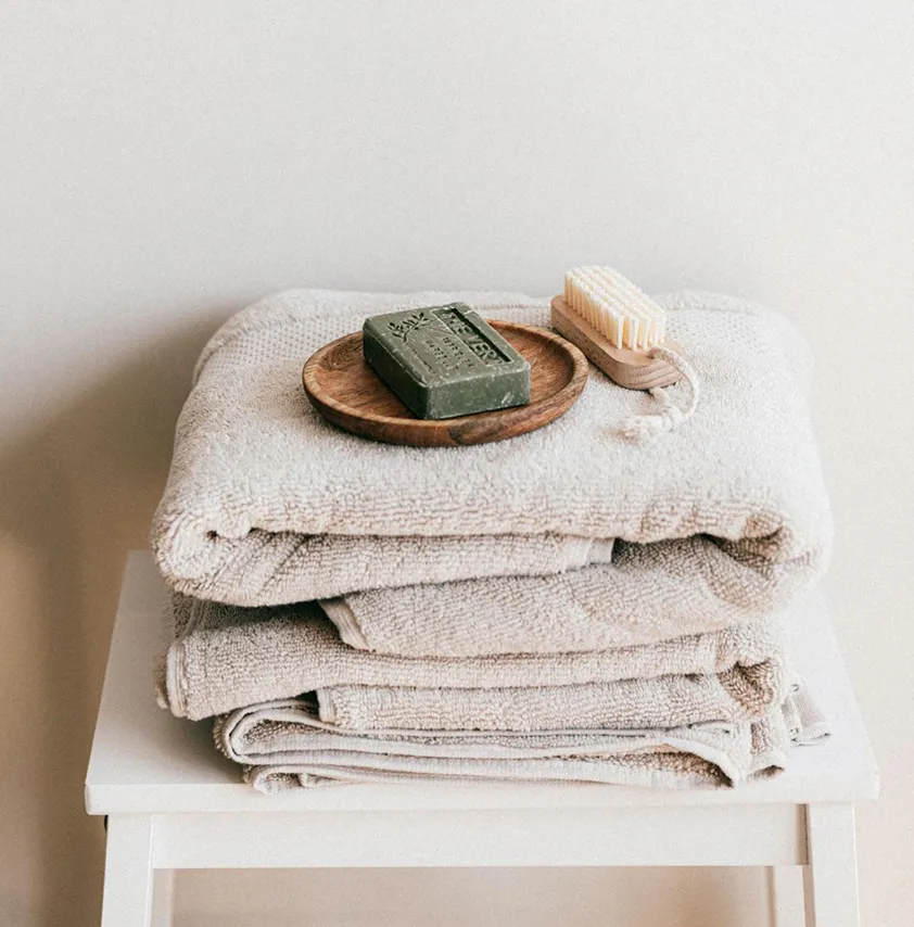 Stack of beige towels on a white stool with a wooden soap dish holding a green soap bar and a small wooden brush on top.