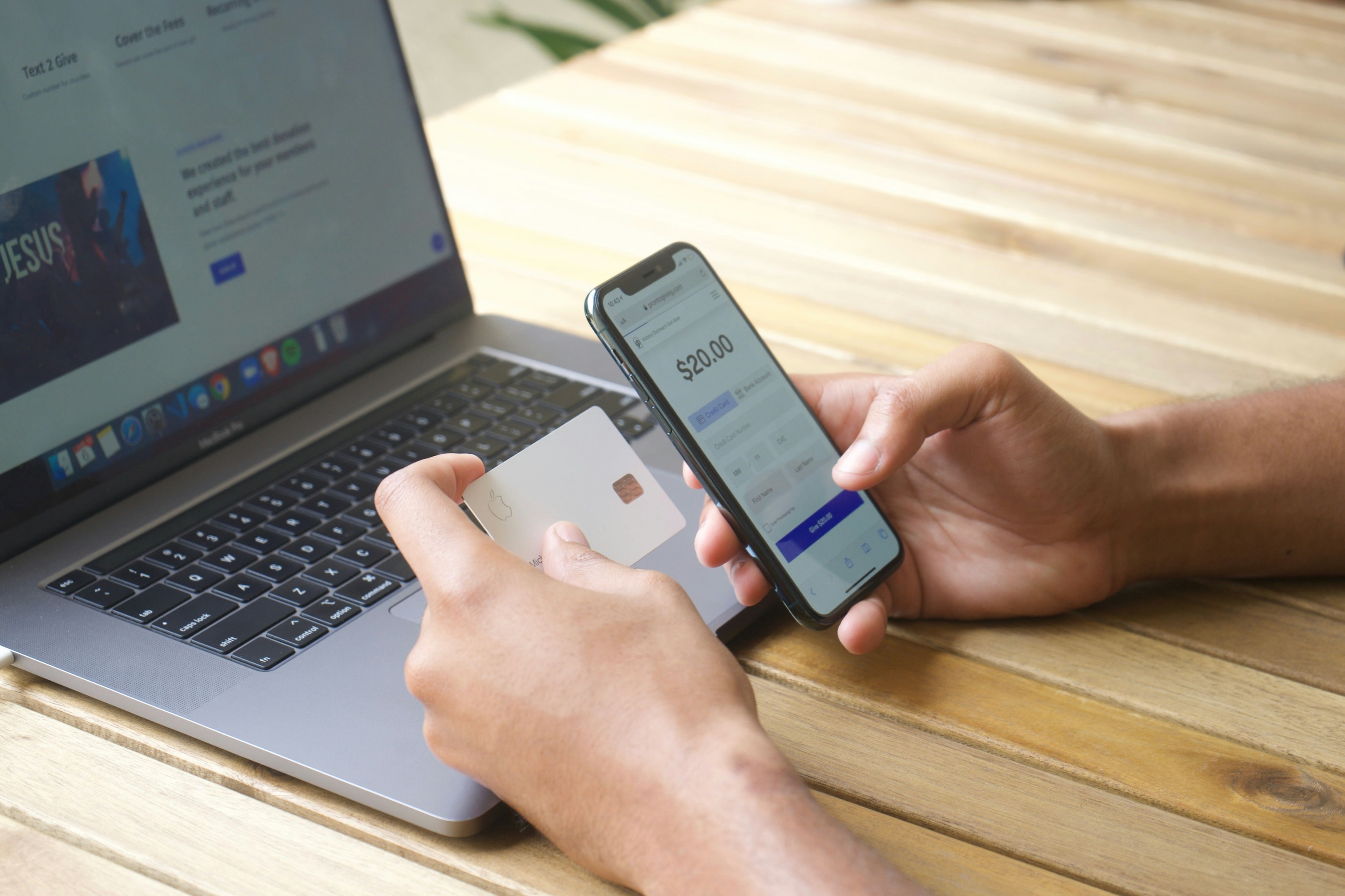 A photo of a man looking at a credit card and his phone, which shows a payment of $20. His laptop is open to a website for a Christian ministry.