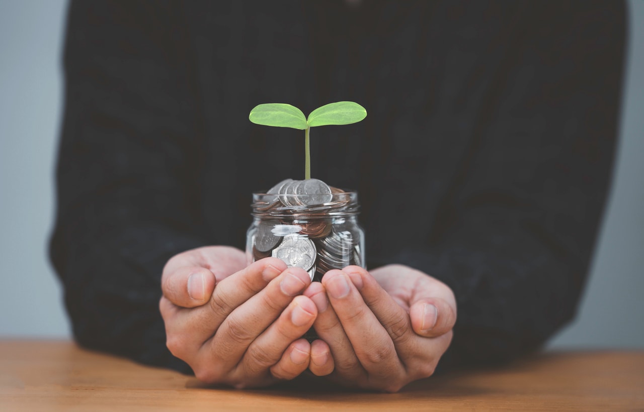A photo of a person holding a jar of coins with a small green plant growing out of the top of the jar.