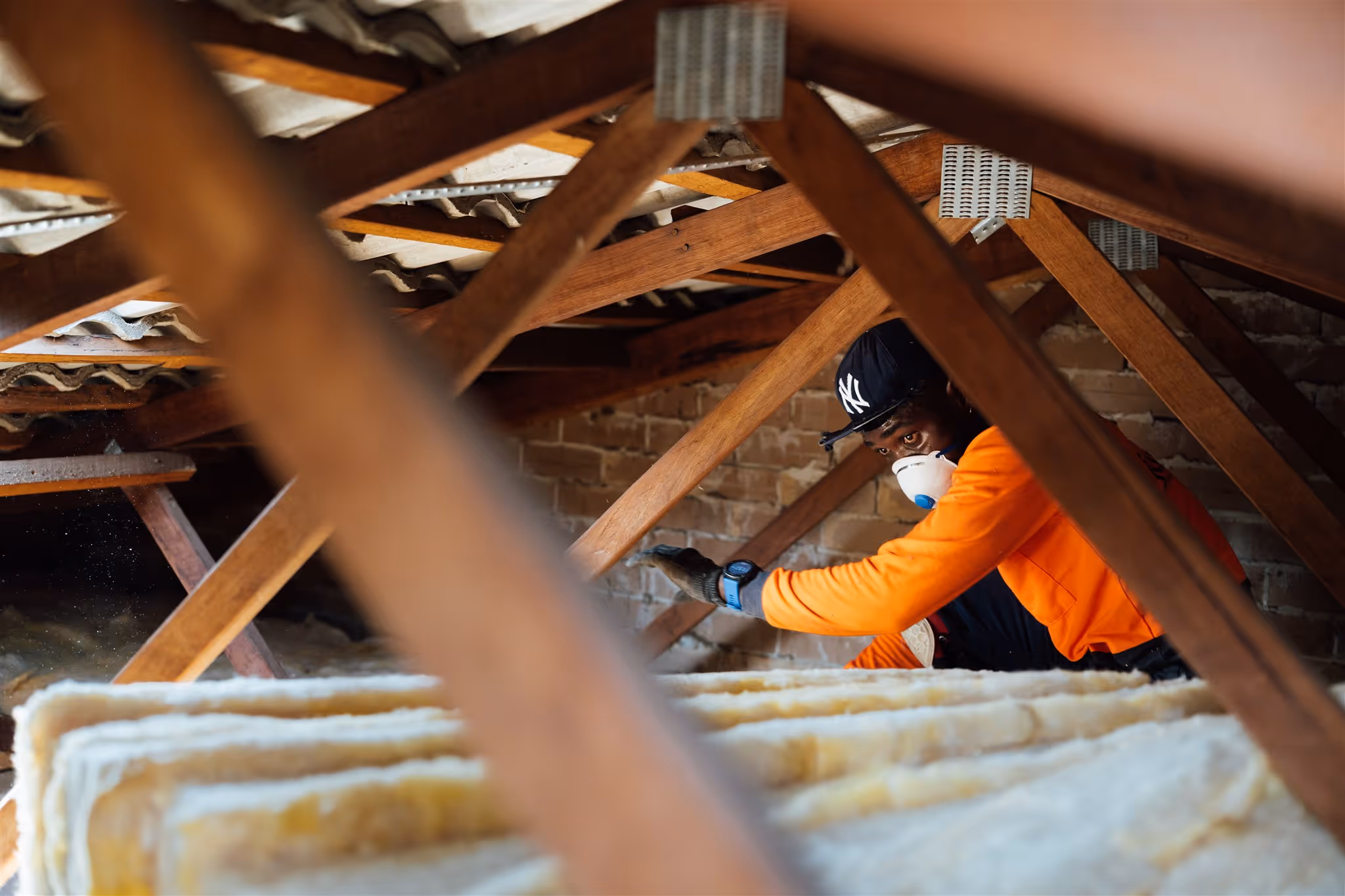 Installer in hi-vis and a dust mask laying insulation batts in a roof space between timber trusses