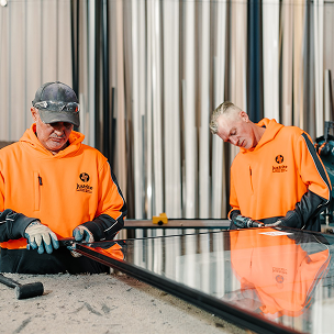 Two workers in hi-vis fitting a frame to a large glass panel in a window workshop