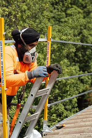 Roof worker in hi-vis, respirator and harness climbing a ladder on scaffolding beside a tiled roof