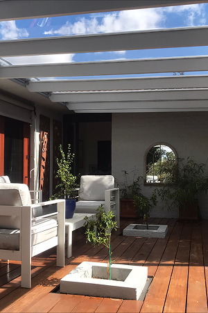 Clear polycarbonate pergola roof over a timber deck with outdoor lounge chairs and potted trees
